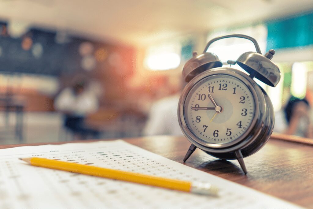 A clock sits on a table next to a standardised literacy and numeracy test while there are teachers completing the LANTITE in the background.