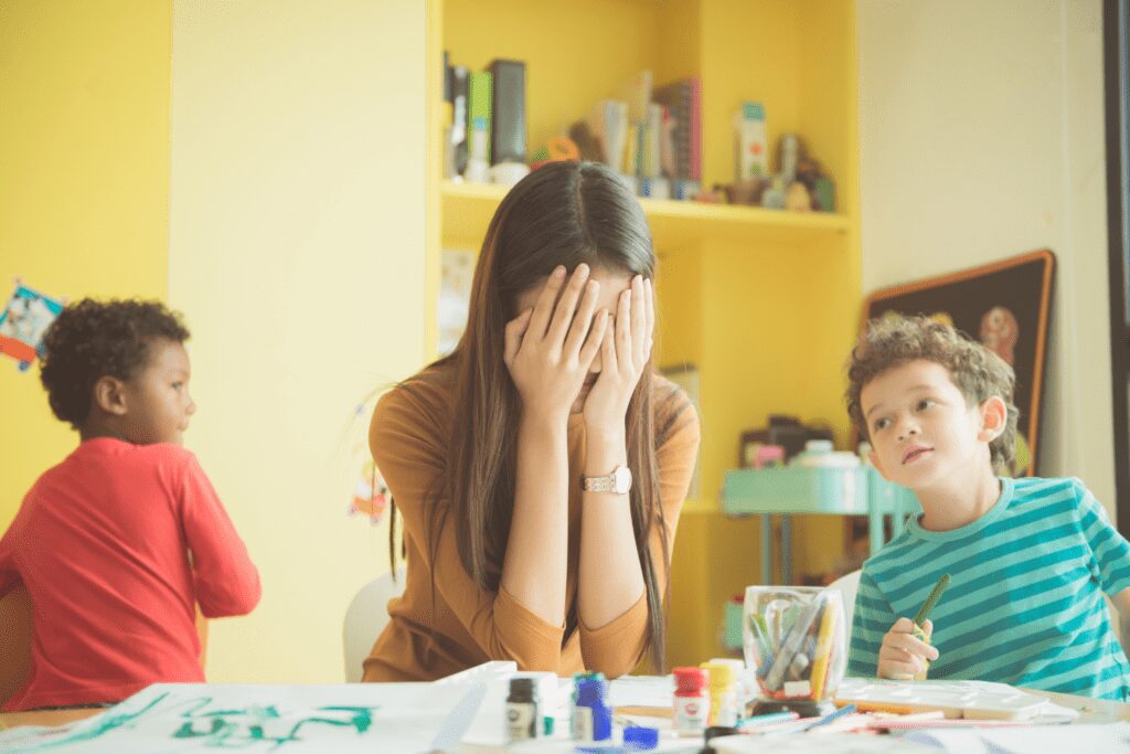 A close up of a teacher with her face in her hands and her elbows on a table, while two of her students are doing craft activities next to her.