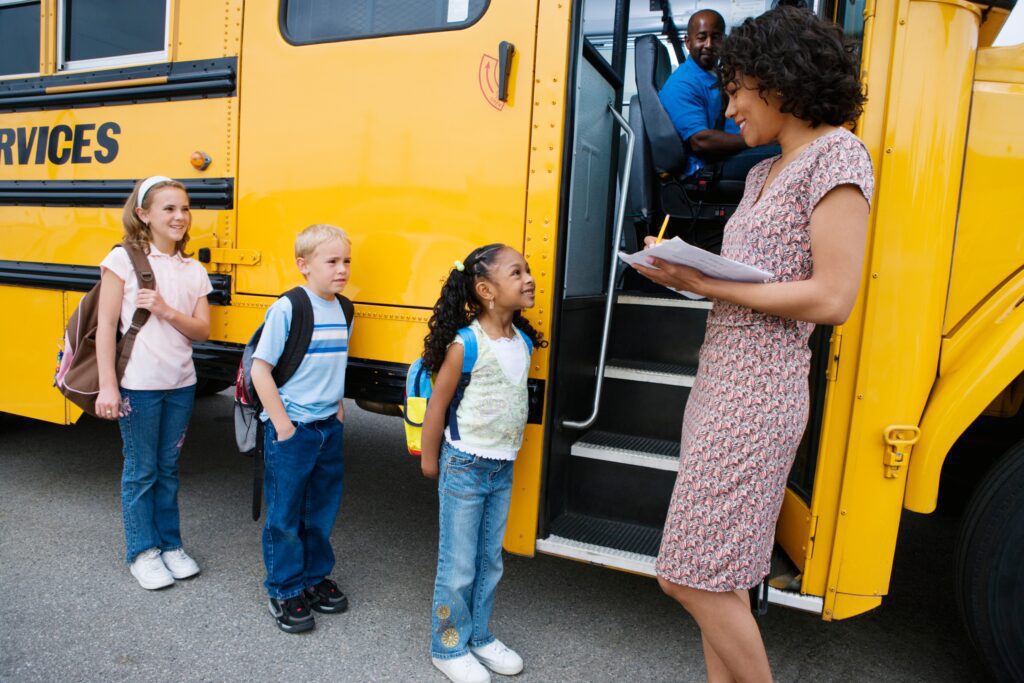 A female teacher is standing in front of the open doors of a school bus. Students are lining up ready to get on, and the teacher is ticking off their names.