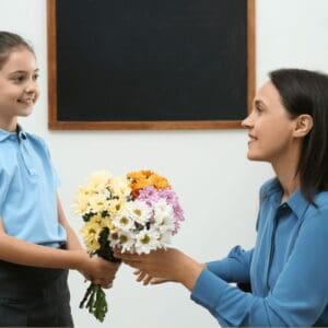 A teacher is kneeling down to give a bunch of flowers to a young student in a uniform. They are both smiling.