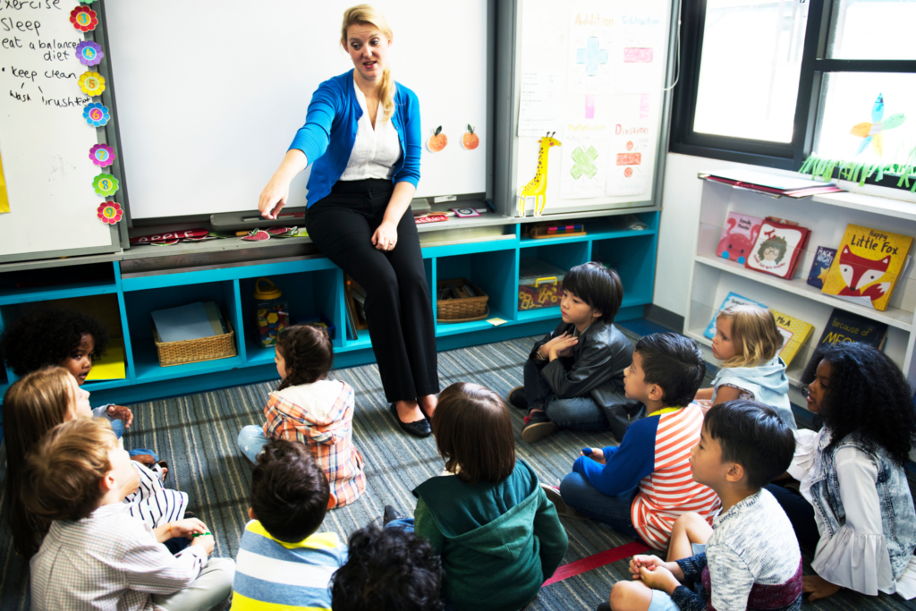 A teacher is standing in front of the whiteboard while her elementary aged students sit on the carpet listening to her teach.