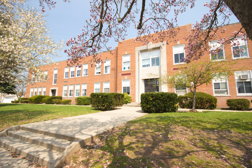 A photo of the front of a public school. The grass is green with some trees and leaves on the ground. The building is made out of bricks and has lots of windows on the facade.