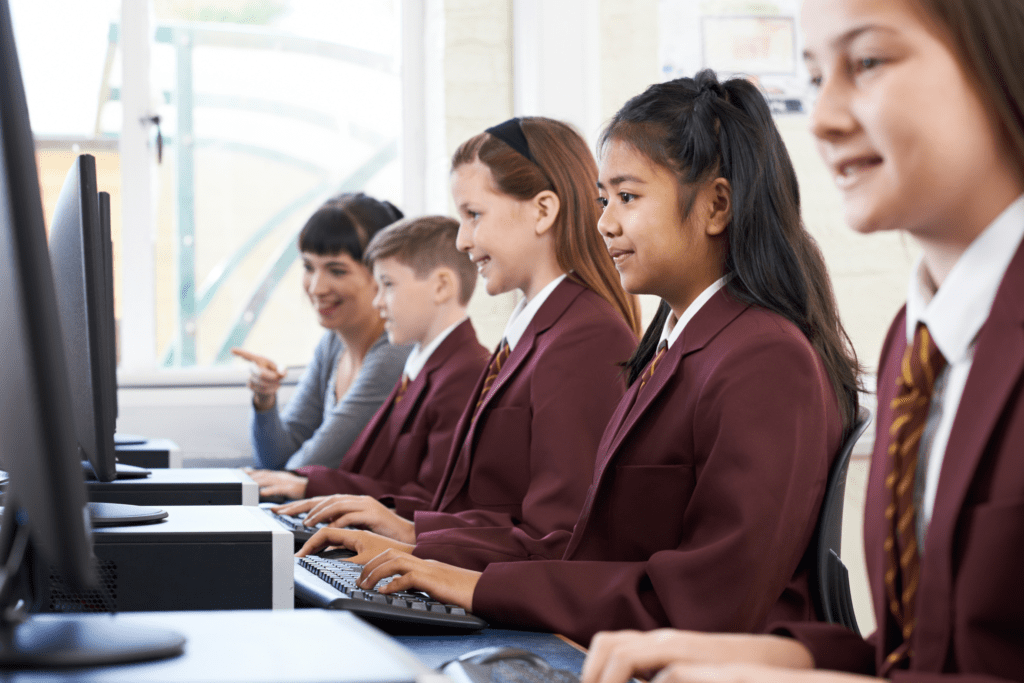 Four private school children in maroon uniforms are sitting in a row working on desktop computers. Their teacher is helping one of them in the background.