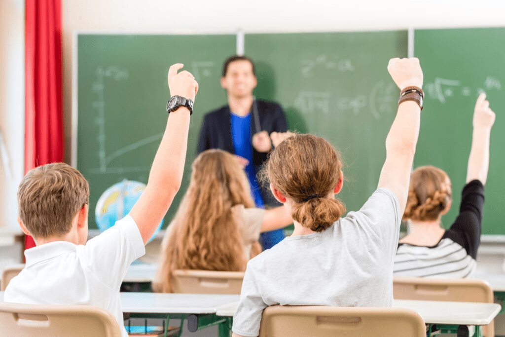 A teacher is standing in front of a blackboard in the background. Rows of students in the foreground all have their hands up to answer a question.