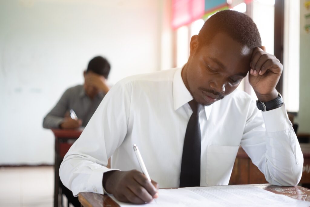 A young pre-service teacher is sitting a literacy test. He is sitting at a desk with his head in one hand. Another teacher is sitting in a desk in the background in a very similar pose.
