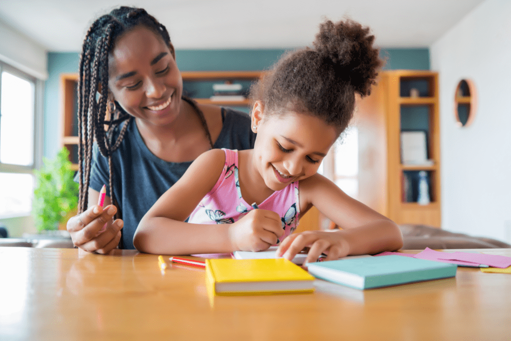 A homeschool student and her mum are smiling as they are both working at a desk writing in small multicoloured notebooks.