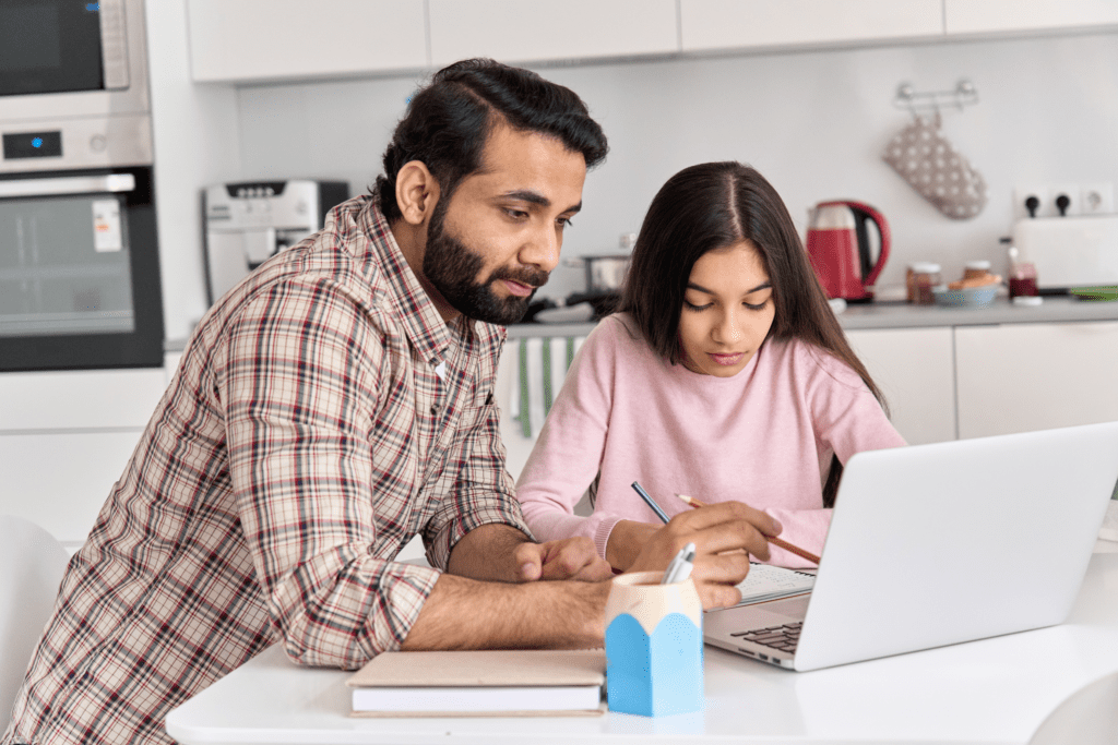 A homeschool student is being helped by her father. They are sitting at the kitchen counter working together on a laptop.