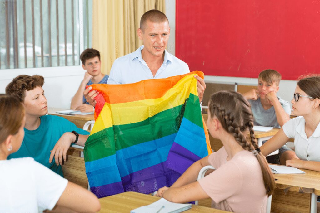 A male teacher is sitting amongst rows of students at wooden desks. He is holding a rainbow flag as he explains it's importance to LGBTQ+ people.