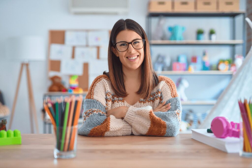 A young female teacher is sitting at her desk smiling. She has a pot of coloured pencils in the foreground on the desk and her classroom is in the background. She has her arms crossed and a soft, warm knitted jumper.