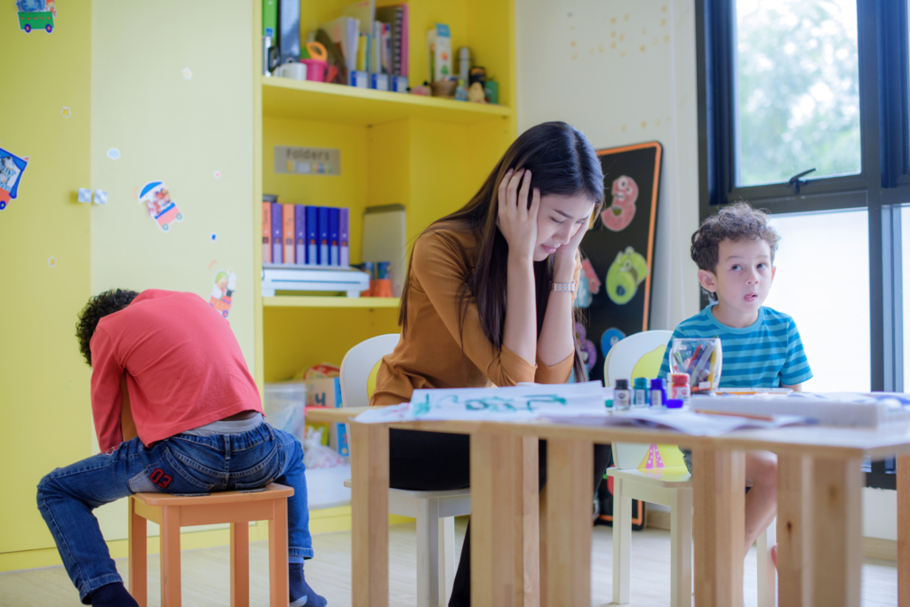 A teacher is sitting at a small table with two of her students doing craft activities. One of the students is sitting backwards on his chair slumped over, and the teacher is resting her head in her hands with her elbows on the table.