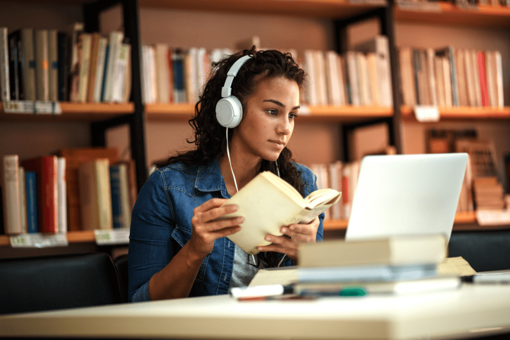 A young college student is studying in a library. She is holding a book and reading off her laptop while listening to music.