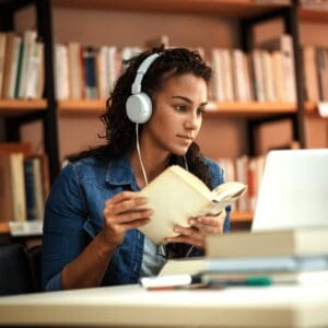 A woman wearing headphones reading a book in a library.
