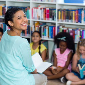 A highly accomplished teacher is sitting in her classroom with her students. They are all sitting on the floor, and the teacher is holding an open book to read to the students. The teacher turns her head around to smile at the camera.