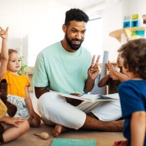 A teacher is sitting on the floor with his young students. He is reading from a picture book and holding up two fingers while looking at one of the students. All of the students are looking at him, some with their hands up.