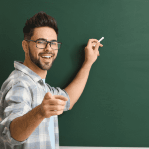 A young male teacher is smiling and pointing at the camera with one hand. In his other hand, he is holding a piece of chalk, poised to start writing on the blackboard behind him.
