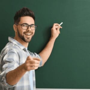 A young male highly accomplished teacher is smiling and pointing at the camera with one hand. In his other hand, he is holding a piece of chalk, poised to start writing on the blackboard behind him.