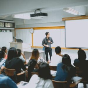 A teacher is standing at the front of his classroom. His class of high school students are sitting at desks in rows. The teacher is turning to look at the projector screen behind him.
