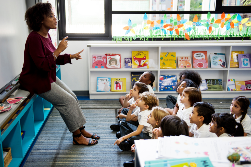 A female teacher is standing and leaning on a shelf at the front of her classroom. Her junior primary class is sitting on the floor in front of her. She is talking and gesturing at them while they listen intently.