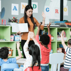 A teacher is standing in front of her young primary school students. There are shelves in the background with an assortment of learning materials. She holds an open book to show the students and holds the other hand up in the air. All her students are sitting in their chairs and holding their hands up.