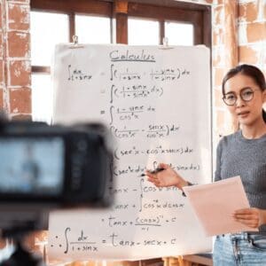 A teacher is standing next to a large piece of paper attached to an easel. She has written maths equations on it, and is explaining them to a camera. She will send the digital recording to her students later.