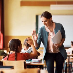 A teacher is walking down the rows of student desks in her classroom. As she walks past one particular student, she is bending down slightly to smile at them and give them a high five.