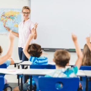 A teacher is standing at the front of her classroom in front of the whiteboard. She is pointing to a country on the world map behind her and looking at the students sitting in rows in front of her. All of her students have their hands up to answer her question.