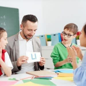 A teacher is sitting at a table in his classroom with his students. They are all around the table, looking at a flashcard that the teacher is holding up. They are all saying the sound "L" that is on the flash card.