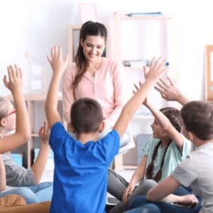 A group of children sitting in a classroom and raising their hands.