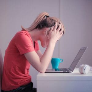 Person in a red shirt sitting at a desk, holding their head in frustration, with a laptop, a blue mug, and headphones nearby.