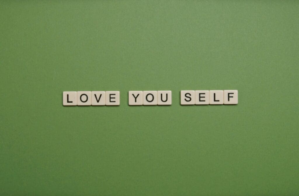Scrabble tiles on a green background spell out "LOVE YOU SELF.