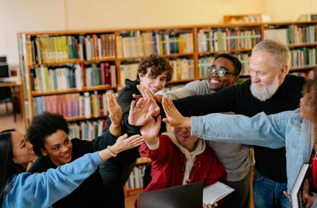 A diverse group of people smiling, standing in a library, and stacking their hands together in celebration or agreement.