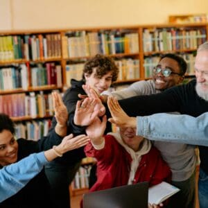 A diverse group of people smiling, standing in a library, and stacking their hands together in celebration or agreement.