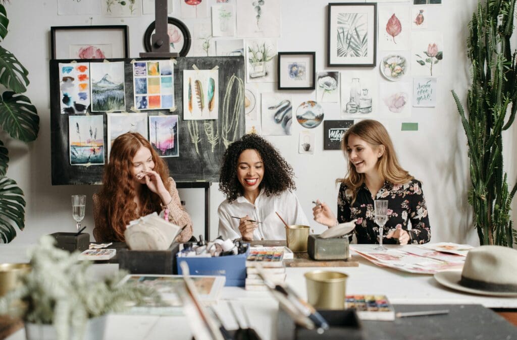 Three women sit at a table, painting and laughing, surrounded by watercolor art and plants in an art studio.