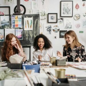 Three women sit at a table, painting and laughing, surrounded by watercolor art and plants in an art studio.